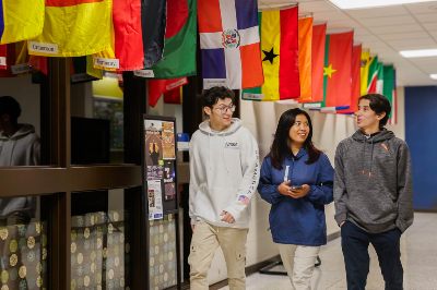 Three students walking down a hall past line of different nation's flags