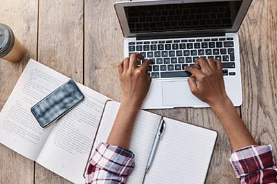 person typing on laptop with open books, pen, phone and coffee