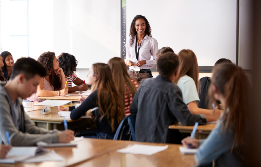 A teacher stands at the front of a classroom, smiling at students who are seated at desks, facing her and taking notes during a lesson.