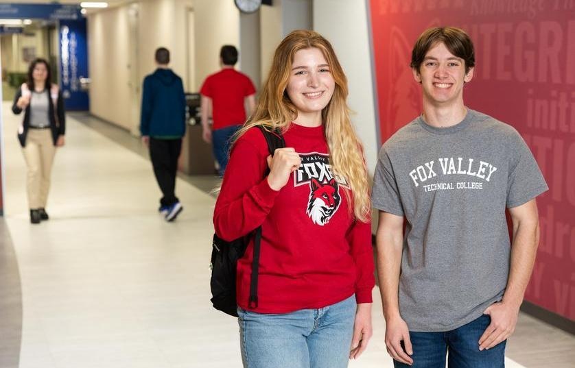 Female and male student standing in brightly lit hallway smiling. The female is wearing a red Foxes long sleeve shirt and the male is wearing a gray Fox Valley shirt. 