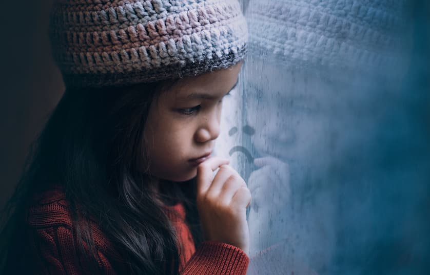 small girl in stocking cap looking sadly out window