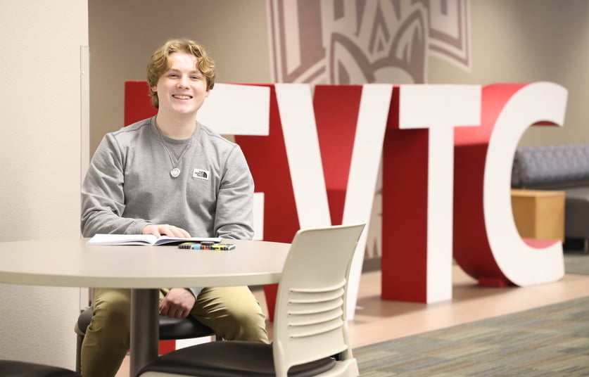 Male student sitting at table smiling in front of large, 3D letters spelling F V T C
