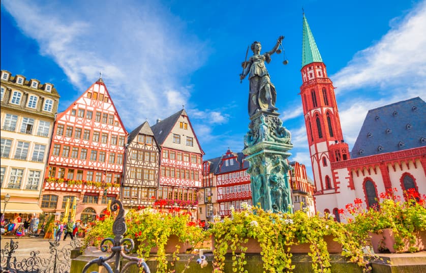 	 A vibrant town square with colorful half-timbered buildings, a statue of Lady Justice holding scales in the center, and a tall church with a green spire under a bright blue sky.