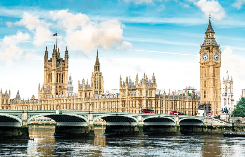 The Palace of Westminster and Big Ben in London, illuminated by sunlight, with a red double-decker bus crossing Westminster Bridge over the River Thames under a partly cloudy sky.