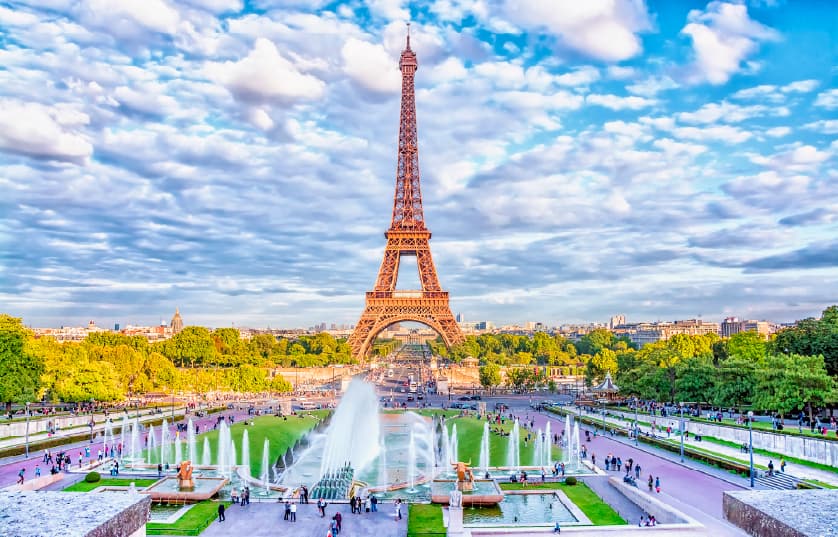 The Eiffel Tower under a partly cloudy blue sky, surrounded by trees and gardens, with fountains and people walking around the Trocadéro Gardens in Paris, France.