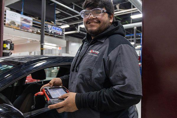 smiling man wearing goggles holding device next to car in automotive shop