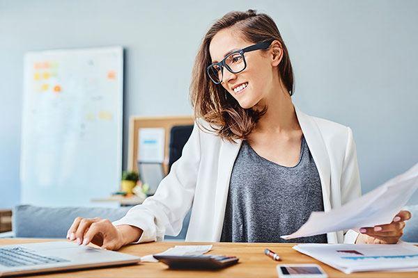 smiling woman in glasses with papers in her hand sitting at desk working on laptop