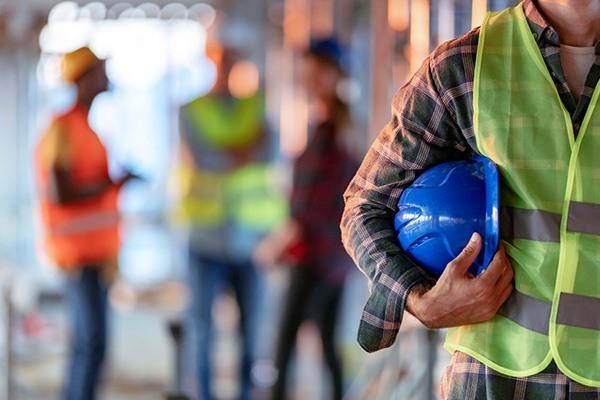 person in flannel and safety vest holding a hardhat with other safety workers in background