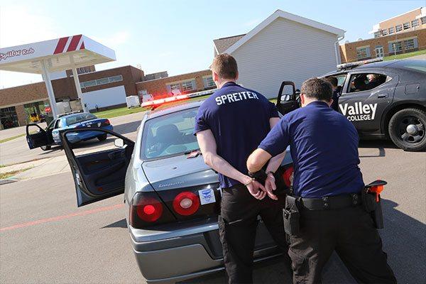 police officer trainee handcuffing person behind training police car with FVTC Public Safety Training Center grounds in background