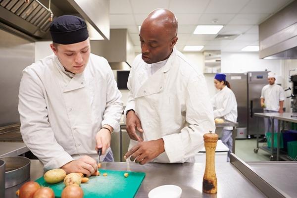 chef teaching culinary student at cutting board with other chefs in kitchen in background