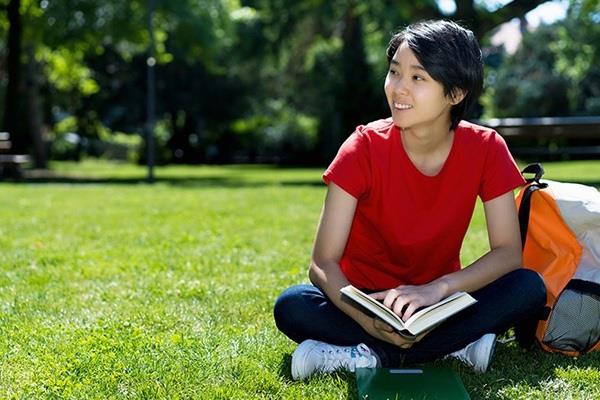 student sitting cross-legged in park with backpack and open book
