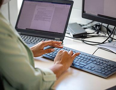 person typing on a keyboard looking toward a laptop and monitor