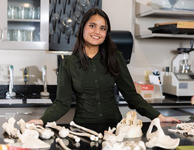 female in black button up shirt standing in a lab behind a counter with bones on it